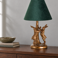 A table lamp with a dark green velvet conical shade and a textured gold-finished base sculpted as two rabbits around a tree trunk, displayed on a wooden sideboard.