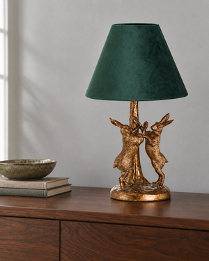 A table lamp with a dark green velvet conical shade and a textured gold-finished base sculpted as two rabbits around a tree trunk, displayed on a wooden sideboard.