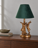 A table lamp with a dark green velvet conical shade and a textured gold-finished base sculpted as two rabbits around a tree trunk, displayed on a wooden sideboard.